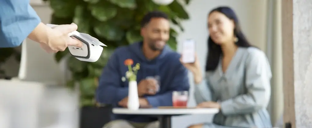 Servicekraft überreicht ein mobiles Gerät an ein Paar, das an einem Tisch sitzt und Getränke genießt, während sie ein Smartphone halten. Der Hintergrund zeigt eine grüne Pflanzenlandschaft, die eine entspannte Atmosphäre schafft.