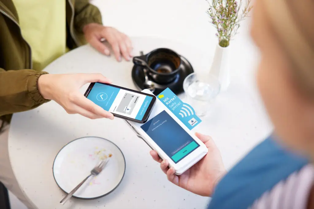 Zwei Personen nutzen ihre Smartphones für eine kontaktlose Zahlung in einem Café. Auf dem Tisch sind ein Teller mit Besteck, eine Tasse und eine Blumenvase sichtbar. Die Bildschirme der Smartphones zeigen Zahlungsanfragen und Informationen zur Transaktion.