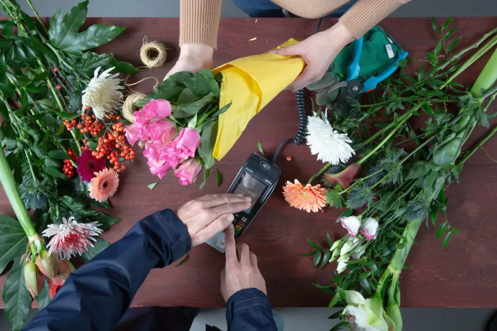 Blumenverkäuferin überreicht einen bunten Blumenstrauß in gelber Verpackung an einen Kunden, während dieser mit einem Kartenlesegerät bezahlt. Der Tisch ist mit verschiedenen frischen Blumen und Pflanzen dekoriert, darunter Rosen, Gerbera und Chrysanthemen.