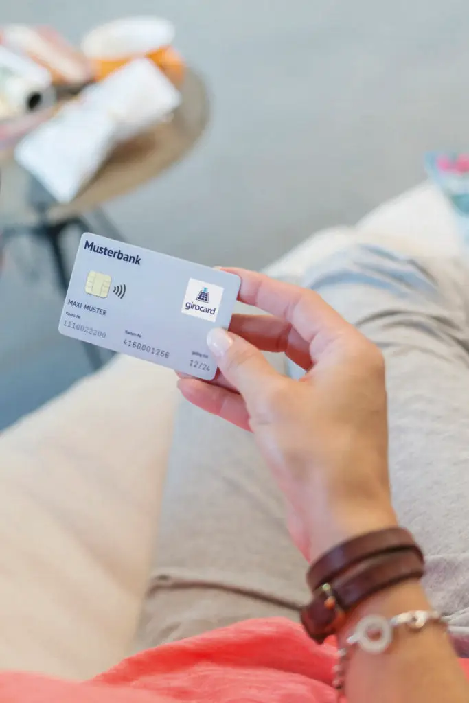 Hand hält eine girocard der Musterbank, die auf einem hellen Sofa liegt, im Hintergrund sind Alltagsgegenstände sichtbar.