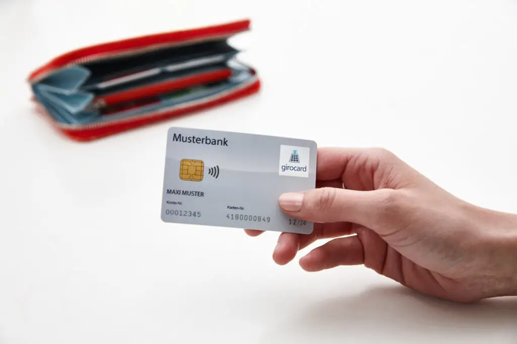 Hand, die eine girocard der Musterbank hält, mit sichtbarem Logo und Karteninformationen, im Hintergrund ein rotes Portemonnaie.