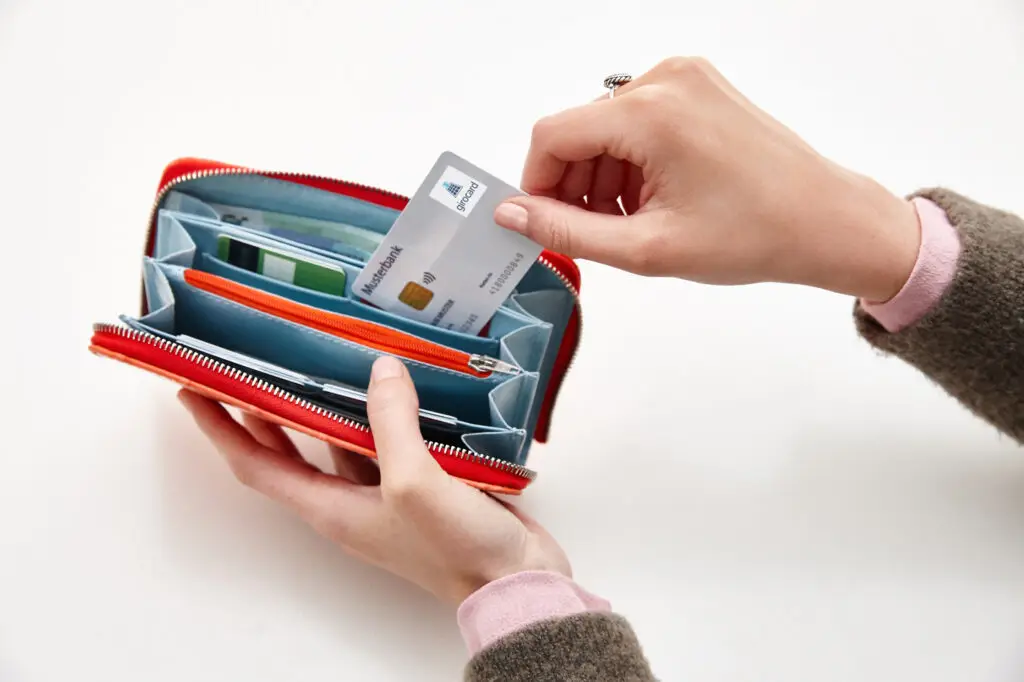 Hände halten ein rotes Portemonnaie, während eine Kreditkarte herausgenommen wird; im Inneren sind mehrere Fächer für Karten und Geldscheine sichtbar, was die praktische Nutzung und Organisation des Geldbeutels zeigt.