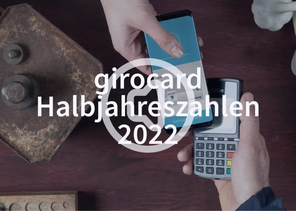 Zwei Hände, eine mit einem Smartphone und die andere mit einem Kartenlesegerät, zeigen eine kontaktlose Zahlung mit girocard im Jahr 2022. Im Hintergrund sind weitere Gegenstände sichtbar, die die Szene ergänzen.