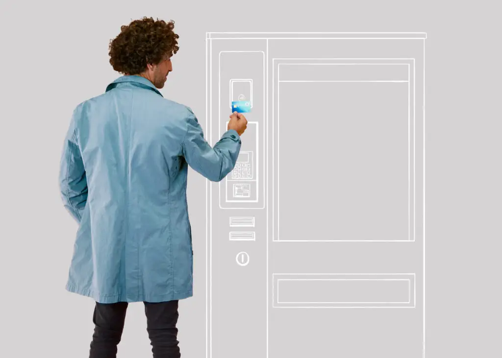 Mann im blauen Mantel nutzt ein kontaktloses Zahlungsmittel an einem Verkaufsautomaten vor einem grauen Hintergrund.