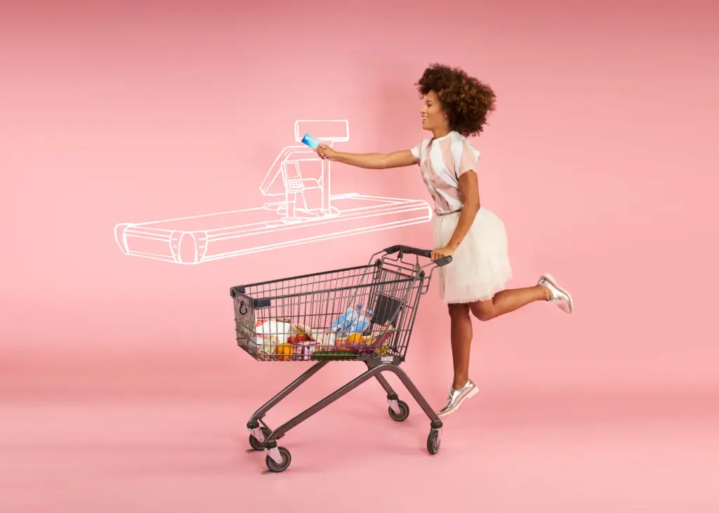 Eine lächelnde Frau mit lockigem Haar steht neben einem Einkaufswagen, der mit Lebensmitteln gefüllt ist, und hält eine Zahlungsmethode in der Hand. Im Hintergrund ist eine skizzierte Kasse auf einem rosa Hintergrund zu sehen.