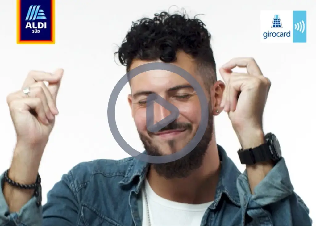 Mann mit lockigem Haar und Bart, der lächelt und die Hände in die Luft hebt, vor einem weißen Hintergrund. Im Bild sind das Logo von ALDI Süd und das Girocard-Symbol zu sehen, wobei ein Play-Button auf das Video hinweist.