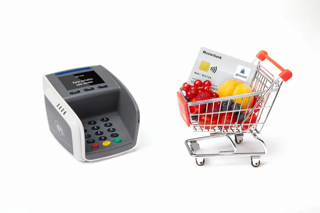 Kassenterminal neben einem Einkaufswagen, der mit frischen Früchten und einer girocard gefüllt ist, symbolisiert modernes Bezahlen im Einzelhandel.