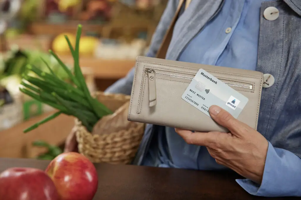 Frau hält eine hellbeige Geldbörse mit einer Bankkarte in der Hand, während sie frische Frühlingszwiebeln in einem Korb trägt, vor einem Marktstand mit Obst und Gemüse im Hintergrund.