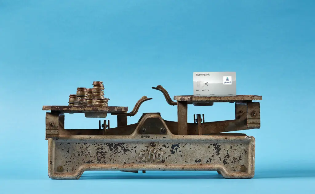 Antike Waage mit Münzstapeln auf der linken Seite und einer Bankkarte auf der rechten Seite, vor blauem Hintergrund, symbolisiert den Wert von Geld und Zahlungsmethoden.