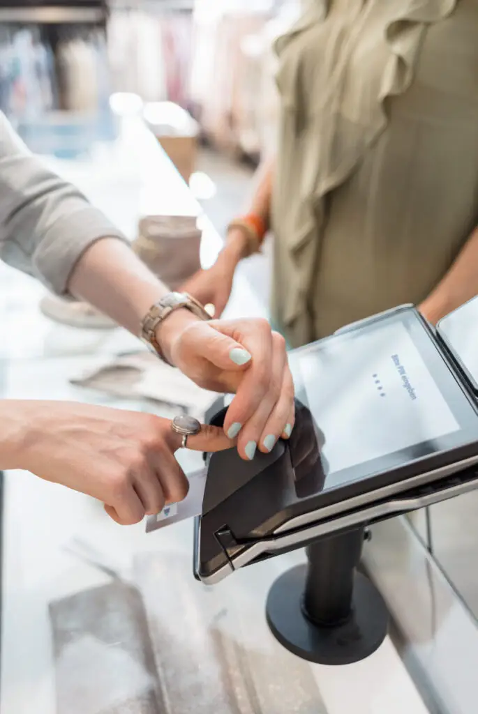 Kundin bezahlt an einem modernen Kassenterminal im Geschäft, während eine Verkäuferin im Hintergrund steht. Die Hände sind auf dem Bildschirm und einer Kreditkarte platziert, was den Zahlungsprozess verdeutlicht.