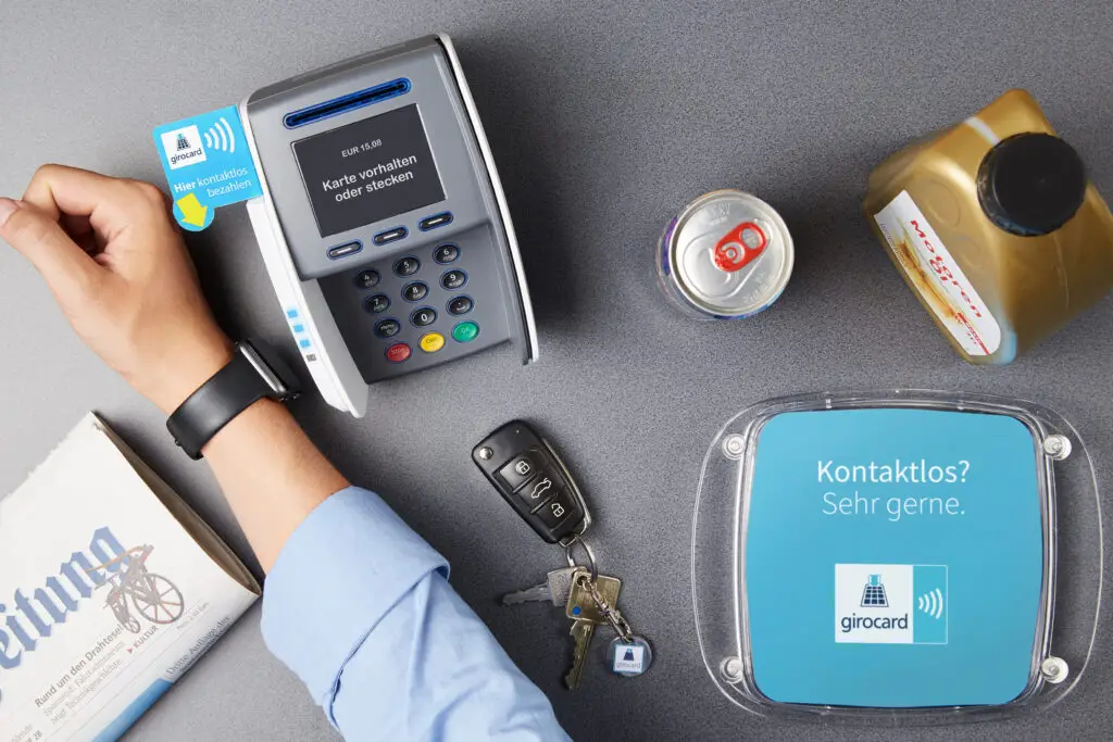 Hand mit Smartwatch, die eine girocard an ein kontaktloses Zahlungsterminal hält, auf einem Tisch mit Getränkedose, einer Flasche und einem Schlüsselbund. Das Terminal zeigt den Betrag von 15,08 EUR an.