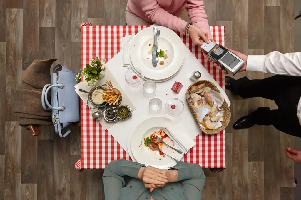 Tischansicht in einem Restaurant mit kariertem Tischdecken, leeren Tellern, einem Kellner, der eine Zahlung entgegennimmt, und einer kleinen Blumendekoration. Im Vordergrund sind eine Hand und eine Handtasche zu sehen, während im Hintergrund ein Korb mit Brot und verschiedene Gewürze auf dem Tisch liegen.