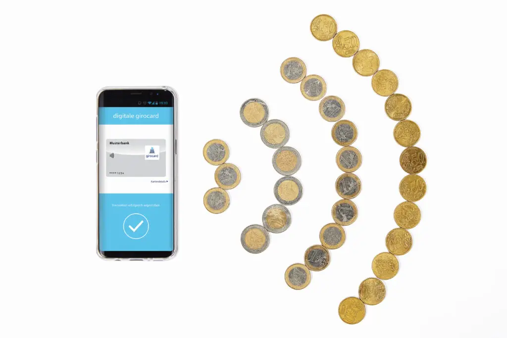 Smartphone mit digitaler girocard-Anzeige neben einer Anordnung von Münzen in verschiedenen Werten, die modernes Bezahlen symbolisiert.