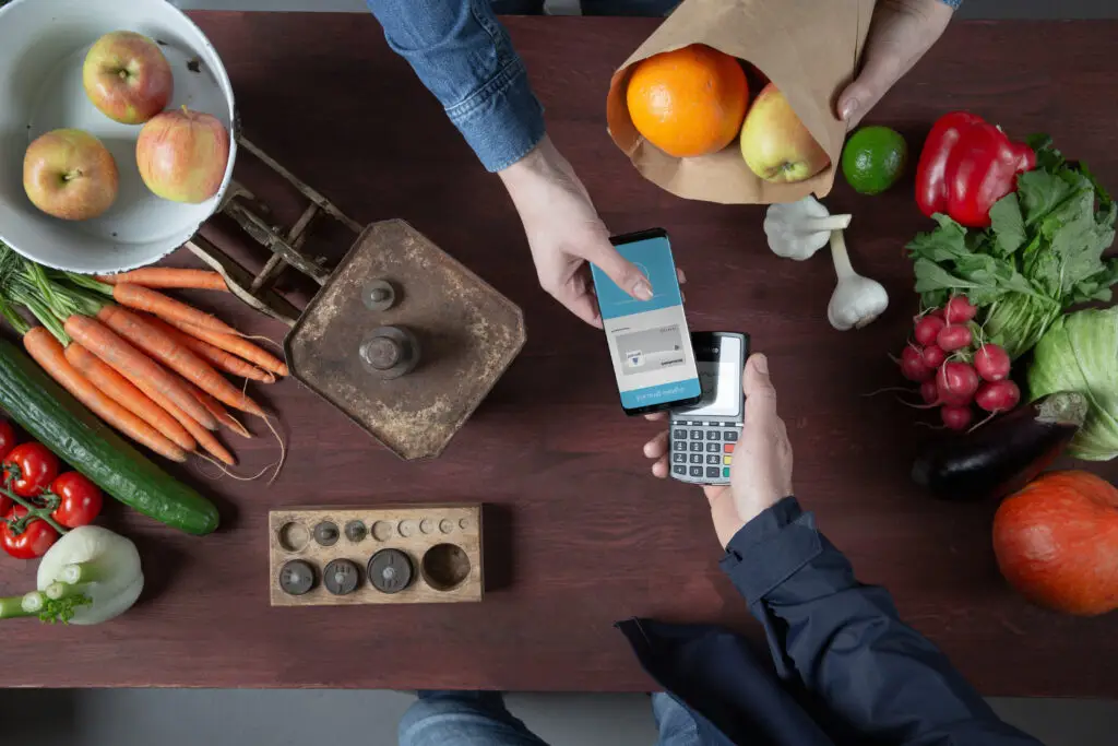 Zahlung mit Smartphone an einem modernen Kartenlesegerät, umgeben von frischem Obst und Gemüse auf einem Holztisch.