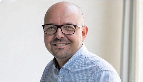 Mann mit Brille und Glatze lächelt freundlich vor einem hellen Hintergrund, trägt ein kariertes Hemd und vermittelt eine positive Ausstrahlung.