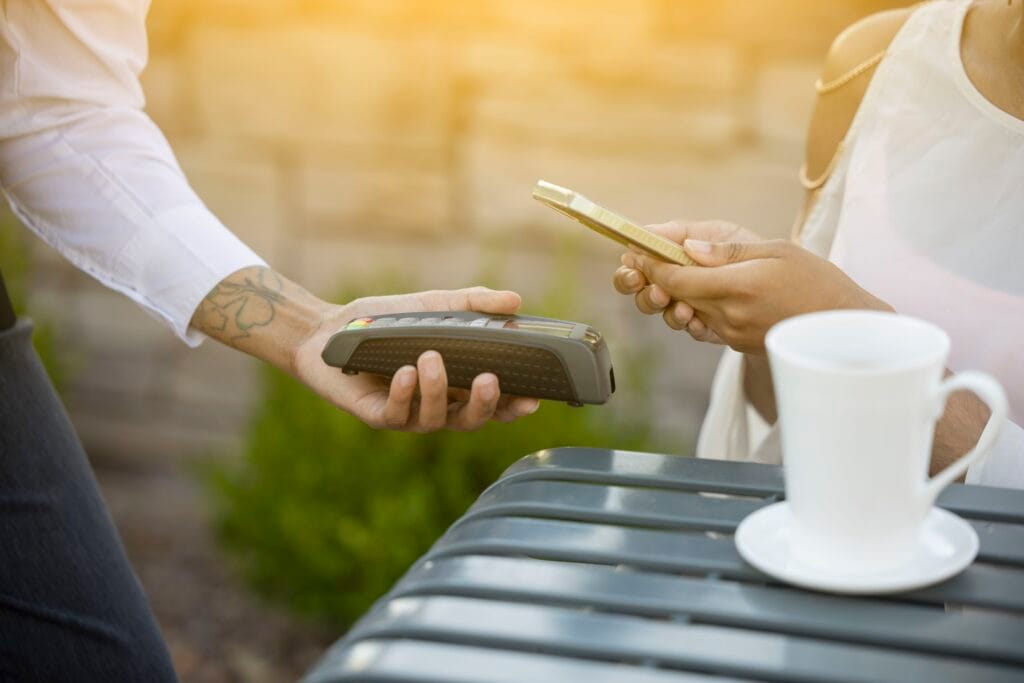 Zahlungsvorgang in einem Café, bei dem ein Kunde sein Smartphone zur Bezahlung nutzt, während ein Mitarbeiter das Kartenlesegerät überreicht. Im Hintergrund ist eine Tasse Kaffee auf dem Tisch sichtbar.