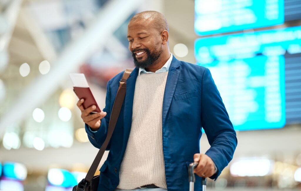 Ein Mann steht mit seinem Reisepass in der Hand am Flughafen. Er lächelt und schaut sein Flugticket an.