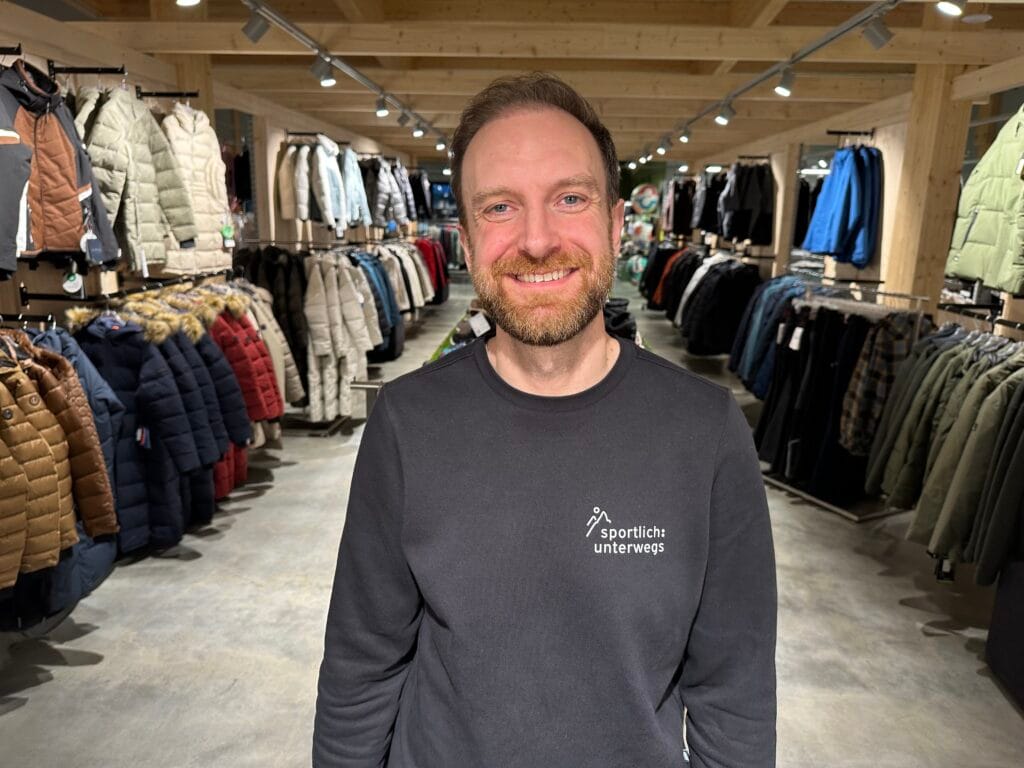 Lächelnder Mann steht in einem Outdoor-Bekleidungsgeschäft zwischen Regalen mit Winterjacken.