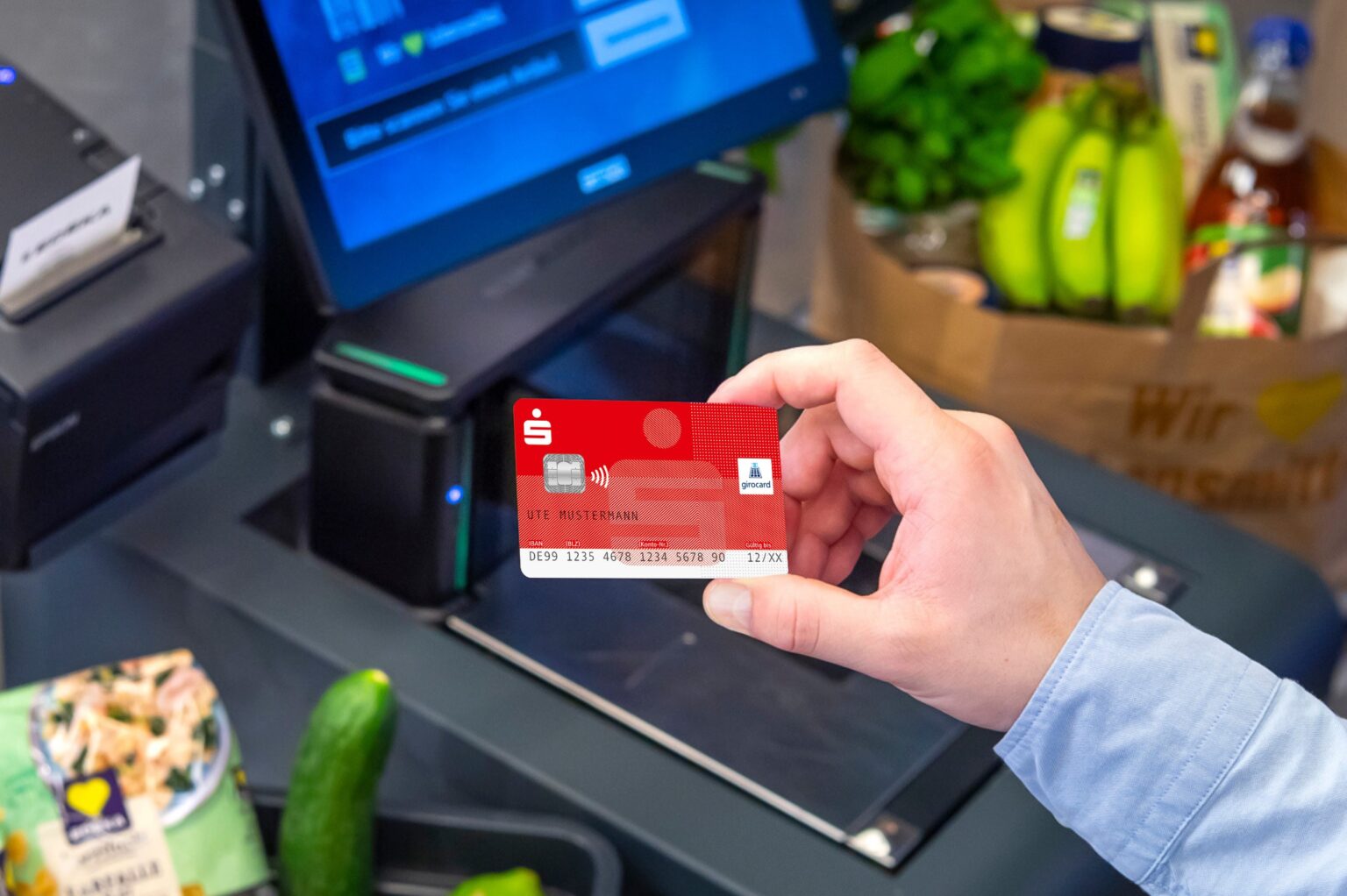 Eine Hand hält in einer Supermarktkassen-Umgebung eine rote girocard der Sparkasse in die Kamera.