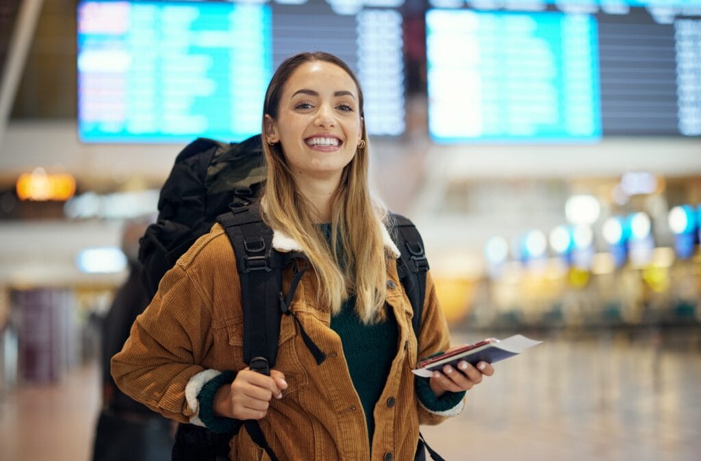 Eine Frau steht mit Reisepass und Smartphone in der Hand am Flughafen und lächelt in die Kamera.
