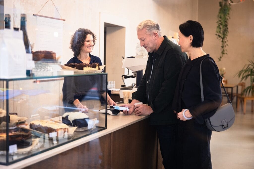 Kundengespräch an der Theke eines Cafés mit einer Verkäuferin, die lächelt, während ein Mann und eine Frau an der Kasse stehen und eine Zahlung tätigen. Im Hintergrund sind verschiedene Kuchen und Getränke sichtbar.