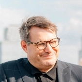 Mann mit Brille und Anzug lächelt vor einer urbanen Kulisse, Hintergrund zeigt verschwommene Hochhäuser.