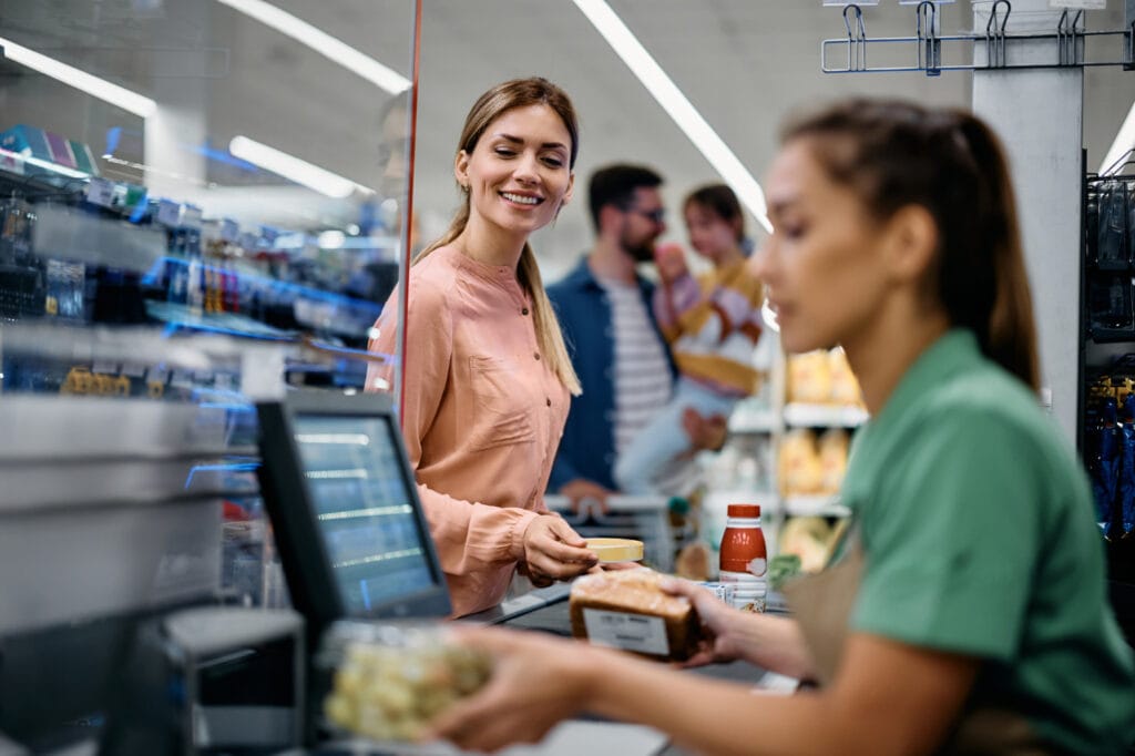 Frau an der Kasse eines Supermarktes, die mit einem Lächeln eine Zahlung tätigt, während eine Kassiererin im grünen Shirt ein Lebensmittel verpackt. Im Hintergrund ist eine Familie zu sehen, die einkauft.