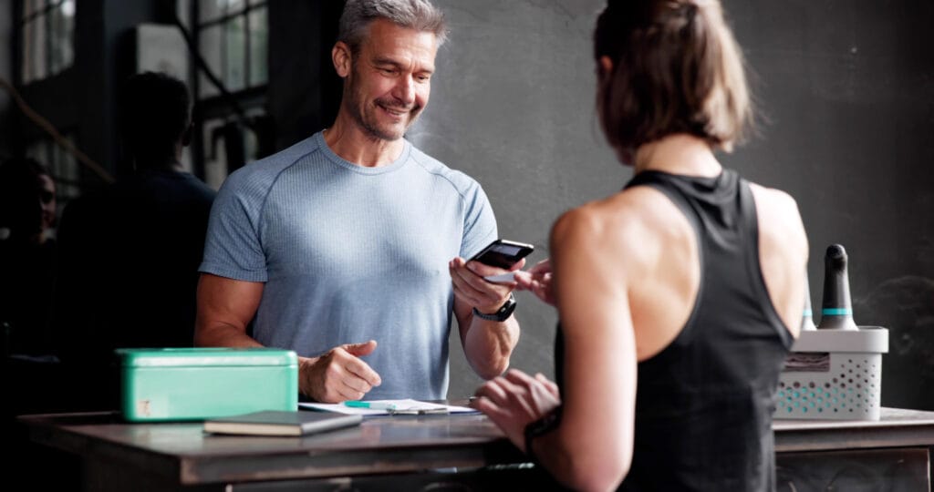 Mann in Sportkleidung lächelt, während er sein Smartphone an eine Frau in einem Sporttop übergibt, die am Empfangstresen steht. Auf dem Tisch sind verschiedene Schreibutensilien und ein grünes Behältnis sichtbar. Die Szene spielt in einem modernen Fitnessstudio oder einer Sporteinrichtung.
