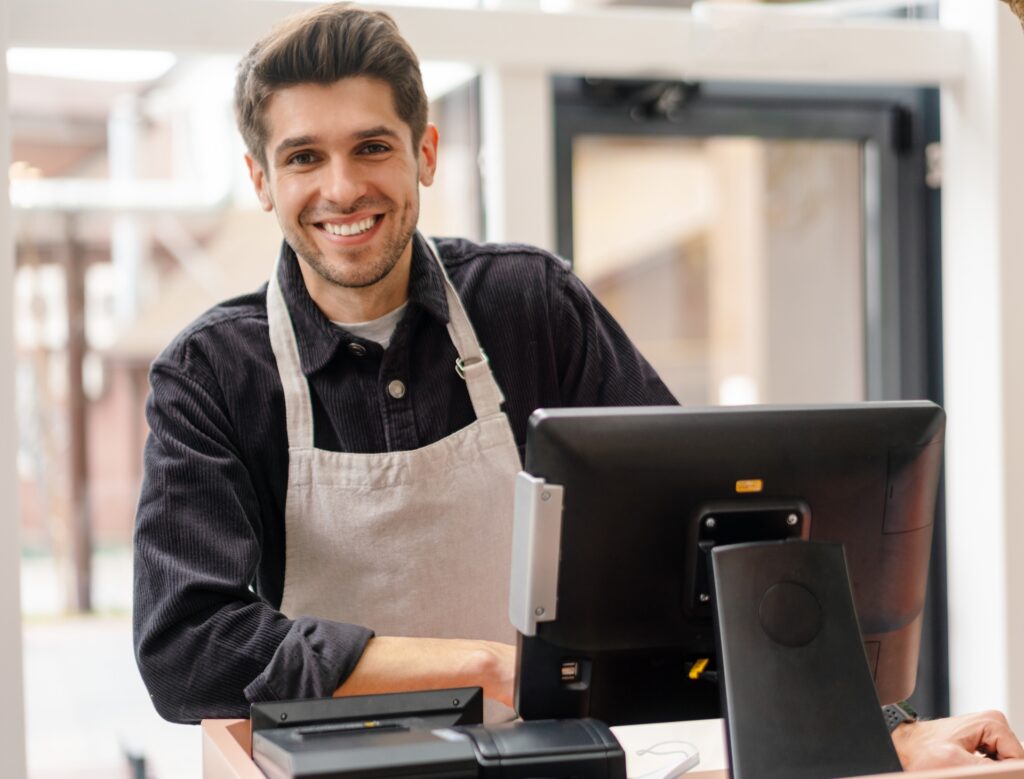 Ein Verkäufer steht an seiner Kasse und lächelt in die Kamera.