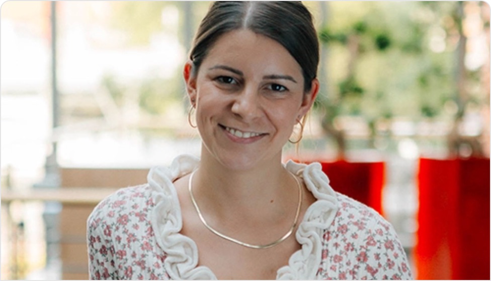 Porträt einer lächelnden Frau mit braunen Haaren, die ein blumiges Oberteil und eine elegante Halskette trägt. Der Hintergrund zeigt unscharfe grüne Pflanzen und dekorative rote Elemente, die eine freundliche Atmosphäre schaffen.